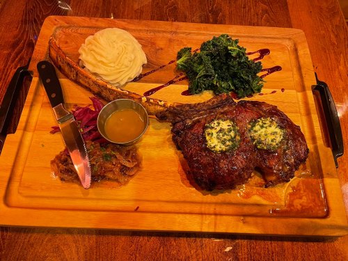 A large ribeye steak with herb butter, mashed potatoes, sautéed greens, and sauce on a wooden platter.