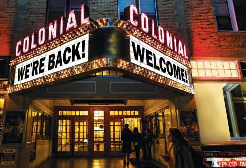 The Colonial Theater entrance lit up at night with a marquee reading "WE'RE BACK! WELCOME!" and people walking by.