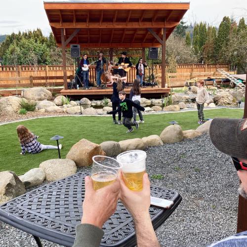 Two people toast with beers in the foreground, while a live band performs on a stage in the background. Kids play nearby.