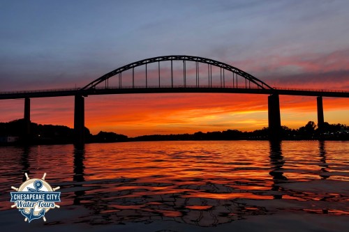 Silhouette of a bridge against a vibrant sunset, reflecting on calm water. Chesapeake City Water Tours logo in the corner.
