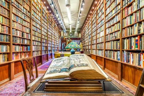 A spacious library filled with tall bookshelves, featuring a large open book on a table and a decorative lamp.