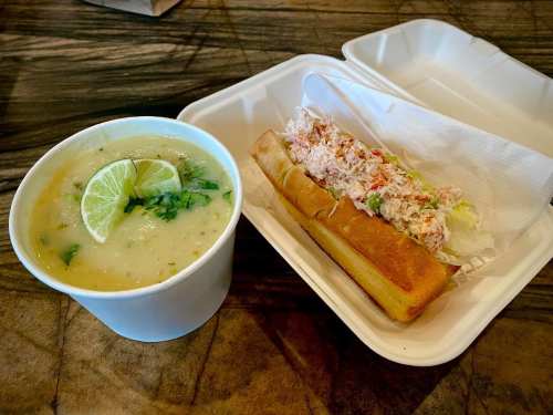 A bowl of soup with lime and cilantro next to a sandwich filled with crab and vegetables in a takeout container.