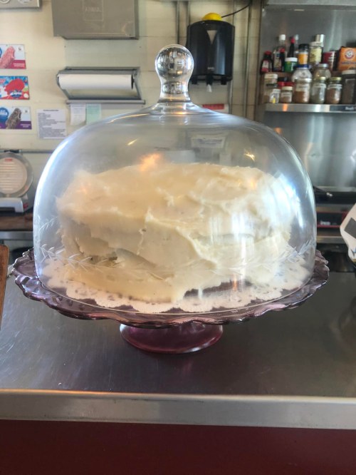 A large cake with white frosting displayed under a glass dome on a decorative stand in a café.