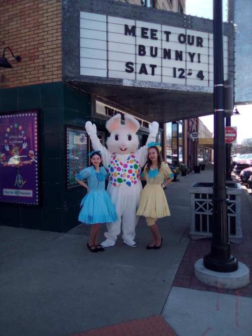 A bunny mascot poses with two women in colorful dresses outside a theater, promoting a special event.