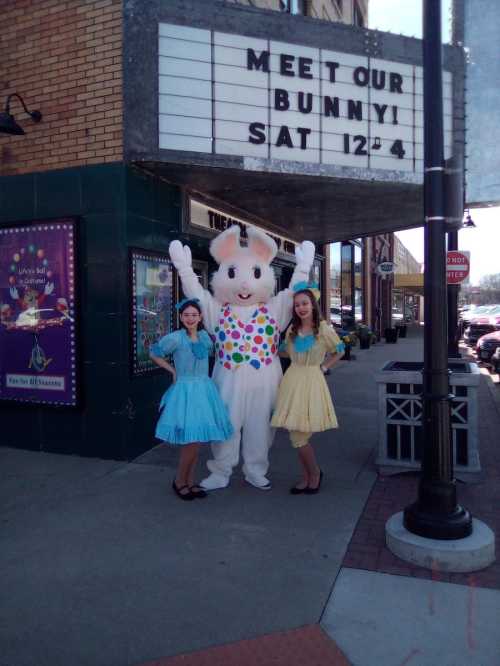 A bunny mascot poses with two women in colorful dresses outside a theater, promoting a special event.