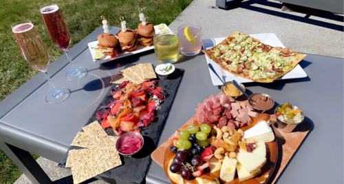 A table with a variety of food: charcuterie boards, sliders, pizza, and drinks in a sunny outdoor setting.