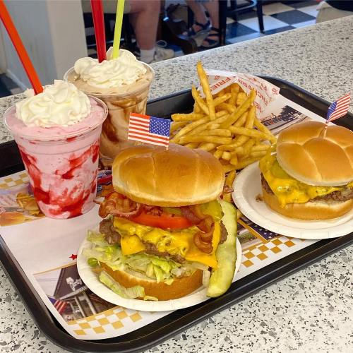 A tray with two burgers topped with bacon and cheese, fries, and two milkshakes with whipped cream and straws.