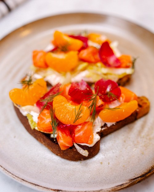 A plate with a slice of dark bread topped with cream cheese, smoked salmon, orange segments, and colorful edible flowers.