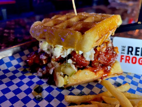 A stacked waffle sandwich filled with fried chicken, drizzled with syrup, served with fries on a blue checkered paper.