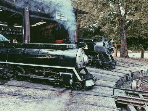 Two vintage steam locomotives at a train station, with smoke rising and trees in the background.
