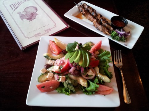 A fresh salad with tomatoes, cucumbers, and feta cheese, alongside a skewer of grilled meat and dipping sauce.