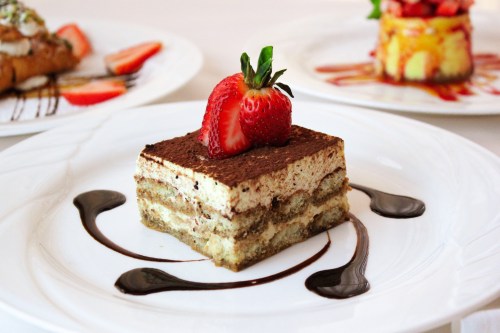 A slice of tiramisu topped with a fresh strawberry, drizzled with chocolate sauce on a white plate.