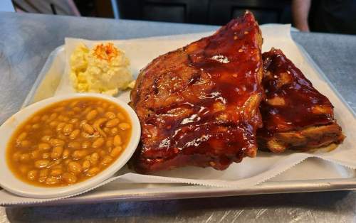 A platter featuring two glazed ribs, a side of baked beans, and creamy potato salad on a metal tray.