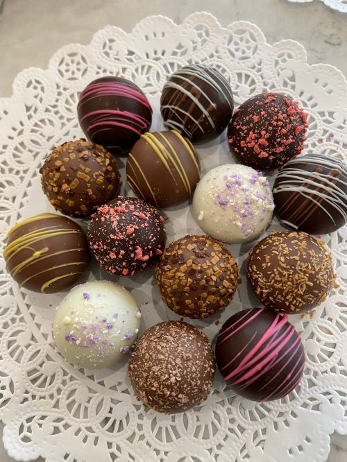 A variety of decorative chocolate truffles arranged on a lace doily, featuring colorful sprinkles and drizzles.