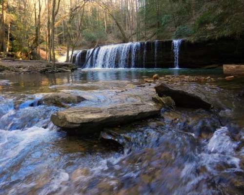A serene waterfall cascades over rocks into a clear stream, surrounded by lush green trees in a tranquil forest setting.