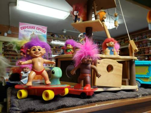 Colorful troll dolls with pink hair skate on toy skateboards near a wooden playset in a vibrant toy store.