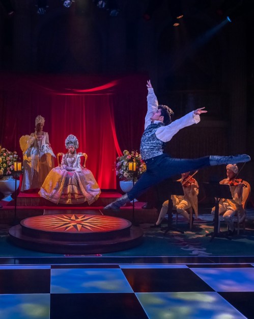 A dancer leaps gracefully on stage, with seated performers in elaborate costumes and a red backdrop.