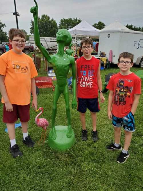 Three boys stand next to a green alien statue at an outdoor market, with a pink flamingo nearby.