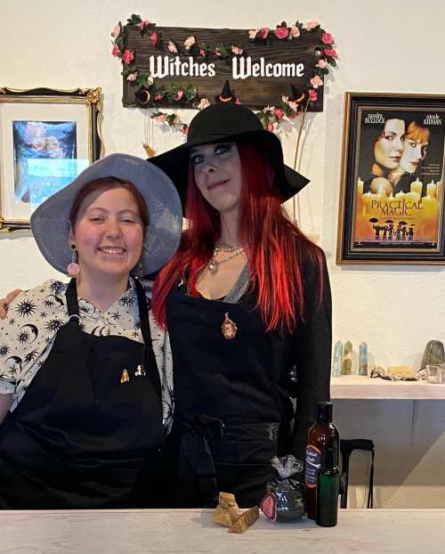 Two women in witch-themed attire stand together, smiling in front of a "Witches Welcome" sign and magical decor.