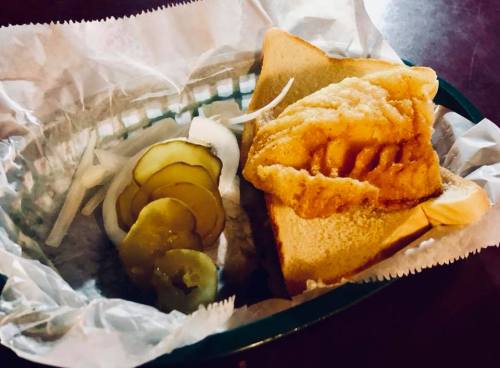A sandwich with fried fish on white bread, served with pickles and onions in a basket lined with paper.