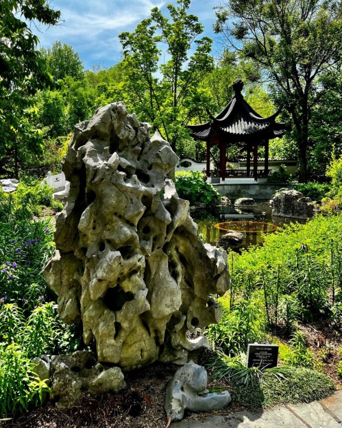 A rocky sculpture in a lush garden, with a traditional gazebo and pond in the background under a blue sky.