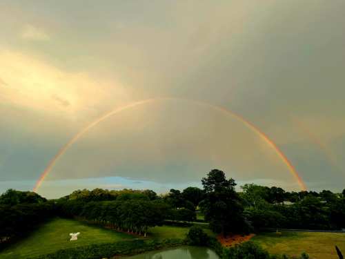 A vibrant rainbow arcs over a lush green landscape with trees and a pond under a cloudy sky.