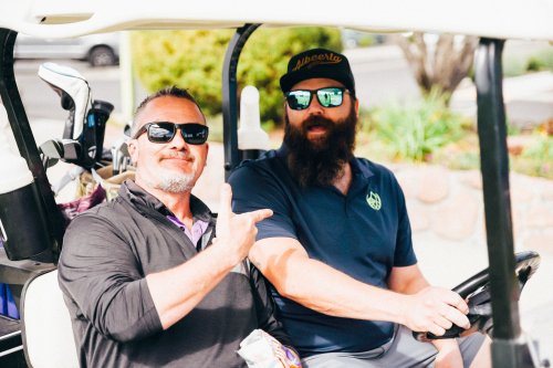 Two men in sunglasses sit in a golf cart, one making a peace sign, with golf clubs visible in the background.