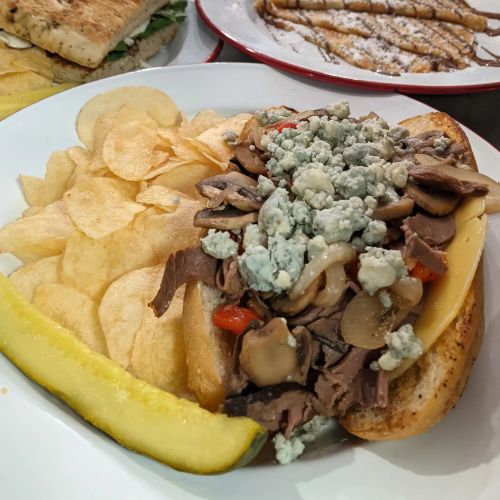 A sandwich filled with beef, mushrooms, and blue cheese, served with potato chips and a pickle on a white plate.