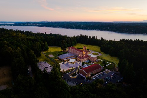 Aerial view of a large building by a lake, surrounded by trees, with a sunset in the background.