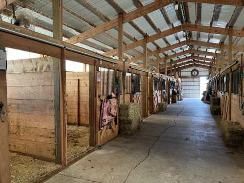 Interior of a barn with wooden stalls, hay bales, and a clear aisle leading to an open door at the far end.