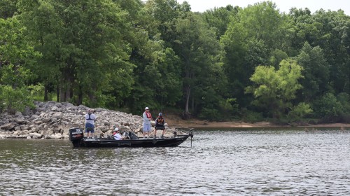 A group of people fishing from a boat on a calm lake, surrounded by lush green trees and rocky shoreline.