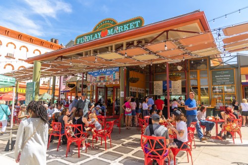 A bustling outdoor market with colorful seating, people dining, and vibrant stalls under a sunny sky.