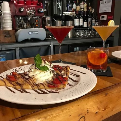 A plate of dessert crepes topped with strawberries, whipped cream, and chocolate drizzle, alongside colorful cocktails.