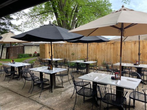 Outdoor dining area with black and beige umbrellas over tables, surrounded by a wooden fence and greenery.