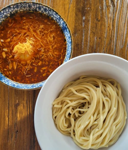 A bowl of noodles next to a bowl of spicy soup topped with cheese and herbs, set on a wooden table.
