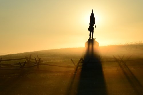 Silhouette of a statue holding a flag against a golden sunrise, with mist and a wooden fence in the foreground.