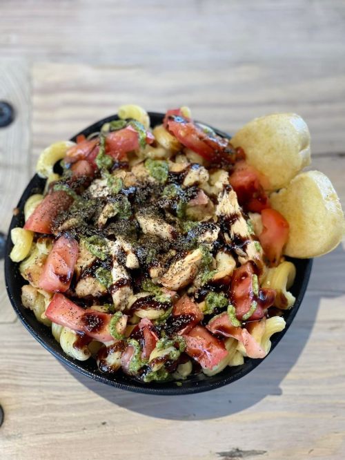 A bowl of pasta topped with grilled chicken, diced tomatoes, and drizzled with sauce, served with two bread rolls.