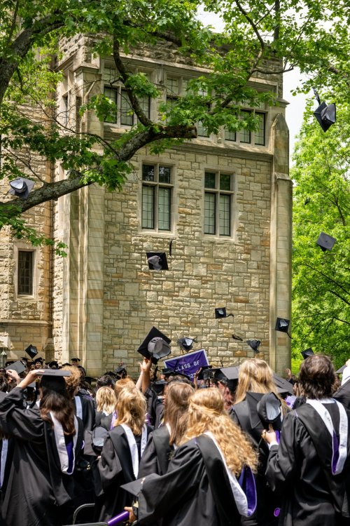 A crowd of graduates in caps and gowns celebrating outdoors, tossing their hats in the air near a stone building.