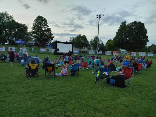 A crowd of people sitting on lawn chairs watching a movie on a large outdoor screen in a park.