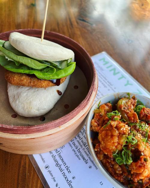 A bao bun with crispy chicken and lettuce beside a bowl of spicy cauliflower and rice, on a wooden table.
