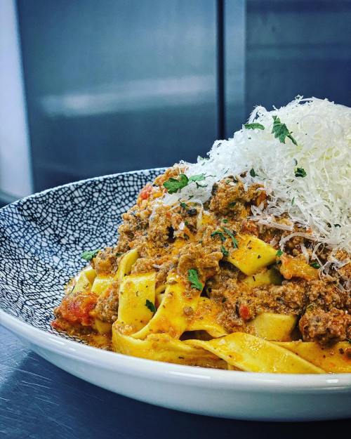 A plate of pappardelle pasta topped with a rich meat sauce and grated cheese, garnished with fresh herbs.