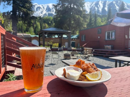 A glass of beer next to a plate of chicken wings and fries, with mountains and trees in the background.