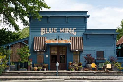 A blue building with "Blue Wing Saloon Cafe" sign, featuring striped awnings and potted plants outside.