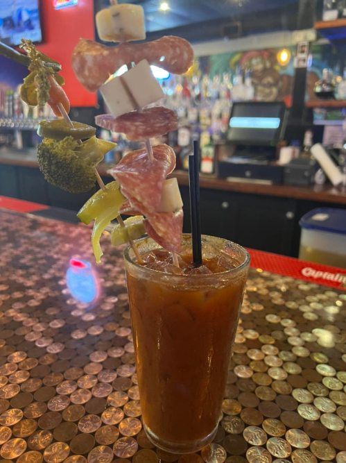 A tall glass of Bloody Mary garnished with skewers of salami, cheese, broccoli, and pickles on a shiny bar counter.