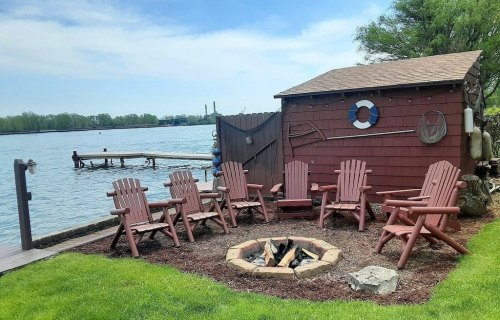 A lakeside scene featuring a fire pit surrounded by wooden chairs, with a shed and dock in the background.