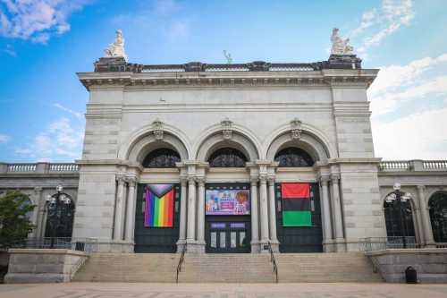 Exterior of a grand building with arched windows, featuring banners in rainbow and red-green colors. Clear blue sky above.