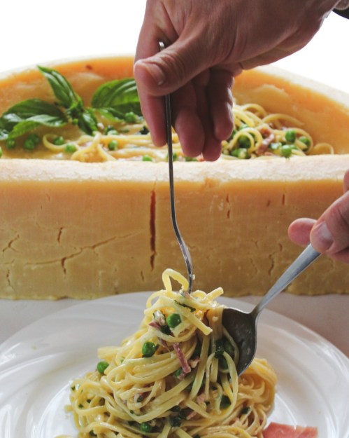 A hand twirls spaghetti with peas and bacon, served from a large cheese wheel on a white plate. Fresh basil garnishes the dish.