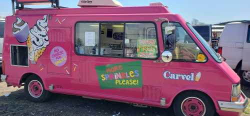 A bright pink ice cream truck with colorful graphics and a sign that says "More Sprinkles Please!" parked at a market.