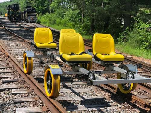 A yellow rail cart with three seats on a railway track, surrounded by greenery and distant trains.