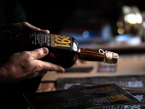 A hand holding a bottle of whiskey with a label reading "169" against a blurred background of menus.